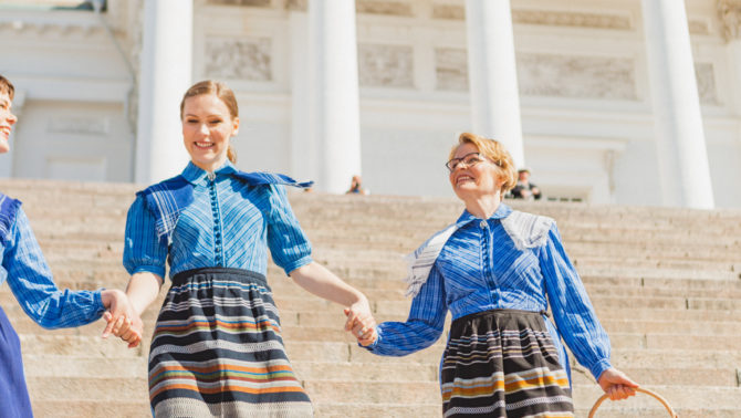 Kolme naista Marttamekoissa kävelevät alas Helsingin Tuomiokirkon portaita ja pitävät toistensa käsistä kiinni. Oikeanpuolimmaisella on kädessään kori ja vasemmanpuolisesta naisesta näkyy vain puolet.