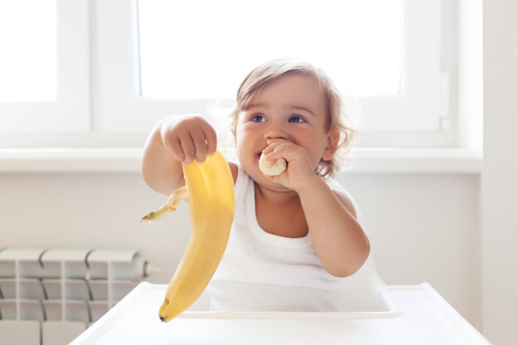 Vauva istuu syöttötuolissa ja pitelee kädessään banaaninkuorta. Hän työntää suuhunsa banaaninpalaa.