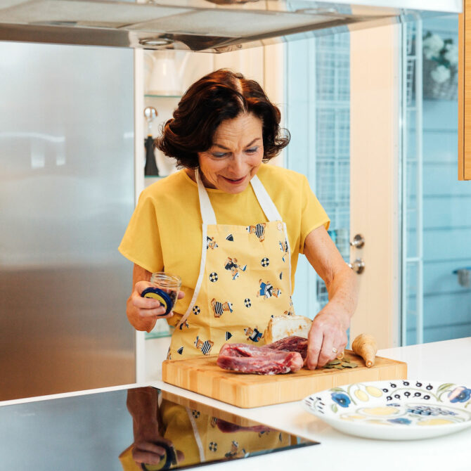 Nainen keltaisessa t-paidassa ja essussa tekee liharuokaa keittiössä.
